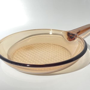 May include: A round, amber-coloured glass skillet with a single handle. The interior base of the pan features a textured, cross-hatched grid pattern, while the rim and handle are a slightly darker shade of brown compared to the translucent body.