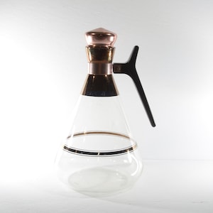 Op de afbeelding: Een glazen karaf met een gouden en zwarte band rond het midden. De karaf heeft een koperkleurig deksel en een zwarte kunststof handvat.