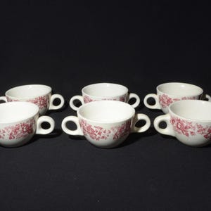 Puede incluir: Seis tazones de sopa de cerámica blanca con diseños florales rojos y dos asas. Los tazones están dispuestos en semicírculo sobre una superficie negra.