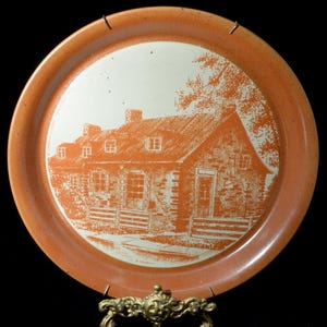 May include: A round, orange-colored serving tray with a white center featuring an orange illustration of a house and surrounding landscape. The tray has a decorative gold-colored handle.