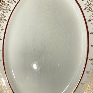 May include: An oval-shaped, off-white serving platter with a cracked glaze. The rim features a dark red border and gold filigree designs. The platter's surface is smooth and slightly reflective, with a subtle pattern of fine cracks.