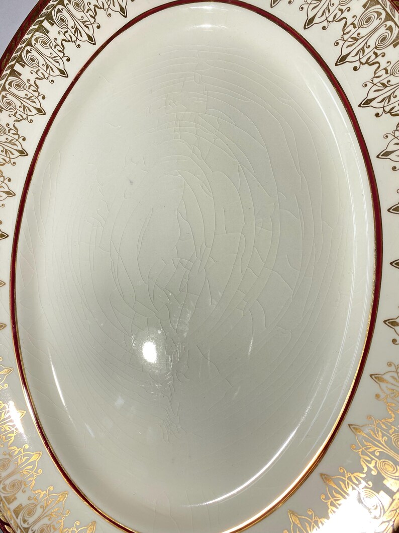 May include: An oval-shaped, off-white serving platter with a cracked glaze. The rim features a dark red border and gold filigree designs. The platter's surface is smooth and slightly reflective, with a subtle pattern of fine cracks.
