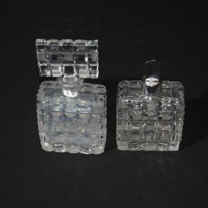 May include: Two clear glass perfume bottles with textured square designs. The bottles have silver tops.