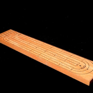 May include: A wooden cribbage board with a natural finish. The board has holes and tracks for scoring, with numbers and markings etched into the wood. The board is rectangular with rounded edges.