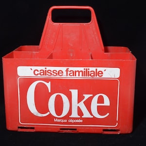May include: A bright red plastic beverage carrier with a central handle and multiple compartments. The front features white text that reads 'caisse familiale', 'Coke', and 'Marque déposée' inside a rectangular border on the front panel.
