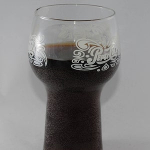 May include: A clear glass with a wide base and a narrow top, filled with a dark brown liquid. The glass has a white Pepsi-Cola logo etched on the side.