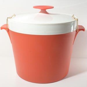 Puede incluir: Una cubeta de hielo vintage de color coral con tapa blanca y un pequeño asa redonda de color coral. La cubeta tiene dos asas laterales y cierres metálicos dorados para asegurar la tapa. La cubeta mide aproximadamente 30 cm de alto.