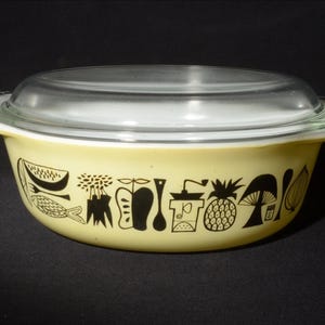 May include: A vintage yellow Pyrex casserole dish with a clear glass lid. The dish features black graphic illustrations of fruits, vegetables, and kitchen tools. The dish has handles on either side.
