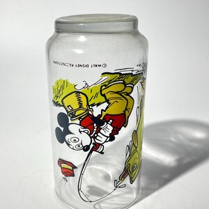 K&ouml;nnte beinhalten: Klares Glas mit grauem Deckel und einer farbenfrohen Illustration von Mickey Mouse beim Angeln. Mickey tr&auml;gt einen gelben Hut und eine rote Hose. Auf der rechten Seite befindet sich ein gelber Fisch. Auf dem Glas steht "&copy; WALT DISNEY PRODUCTIONS".