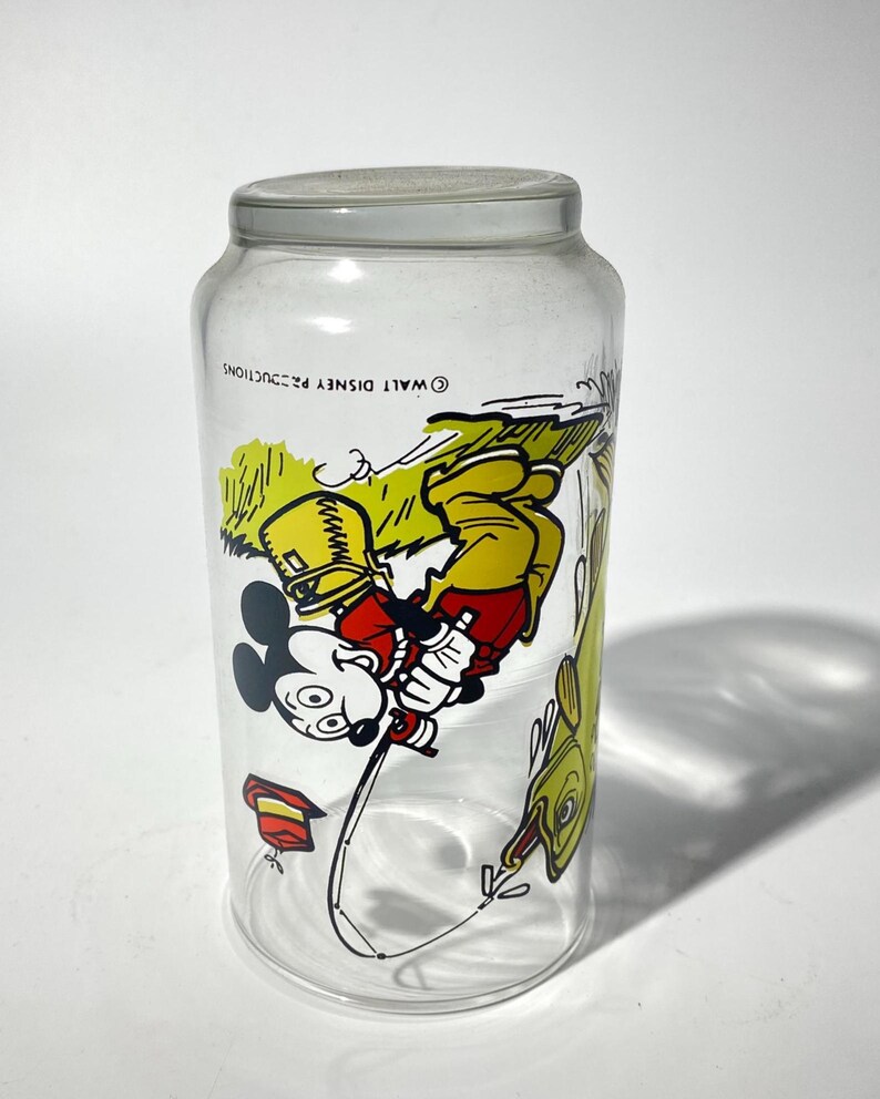 K&ouml;nnte beinhalten: Klares Glas mit grauem Deckel und einer farbenfrohen Illustration von Mickey Mouse beim Angeln. Mickey tr&auml;gt einen gelben Hut und eine rote Hose. Auf der rechten Seite befindet sich ein gelber Fisch. Auf dem Glas steht "&copy; WALT DISNEY PRODUCTIONS".