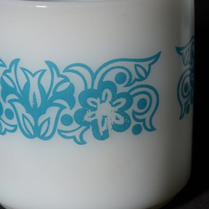 May include: White ceramic mug with a turquoise floral pattern. The mug has a handle and a band of stylized flowers and leaves around the middle. The design is repeated around the mug. The mug is set against a dark background.