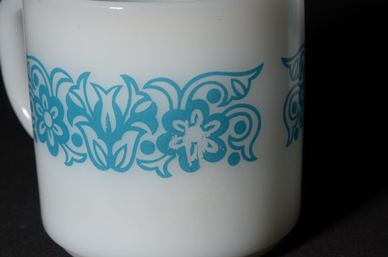 May include: White ceramic mug with a turquoise floral pattern. The mug has a handle and a band of stylized flowers and leaves around the middle. The design is repeated around the mug. The mug is set against a dark background.