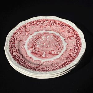 Puede incluir: Una pila de tres platos de cerámica blanca con un patrón de transferencia rojo. El centro de cada plato representa un paisaje con un edificio y figuras, rodeado por un borde ancho de hojas de vid y motivos de enredaderas detallados.