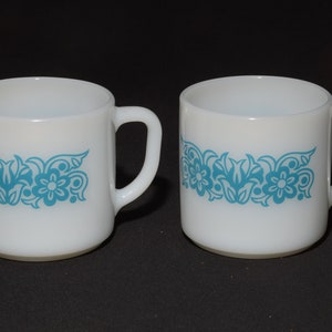 May include: Two white ceramic mugs with a turquoise floral design. Each mug has a handle and a band of turquoise flowers and leaves around the middle. The mugs are set against a black background.