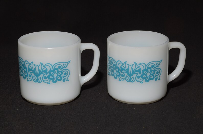 May include: Two white ceramic mugs with a turquoise floral design. Each mug has a handle and a band of turquoise flowers and leaves around the middle. The mugs are set against a black background.