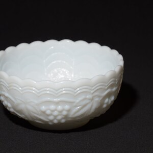 K&ouml;nnte beinhalten: Eine wei&szlig;e, dekorative Schale mit gewelltem Rand und gepr&auml;gtem Trauben- und Blattdesign. Die Schale besteht aus undurchsichtigem Glas und steht auf einer schwarzen Oberfl&auml;che. Die Schale ist ein Vintage-St&uuml;ck.