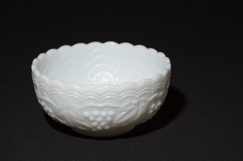 K&ouml;nnte beinhalten: Eine wei&szlig;e, dekorative Schale mit gewelltem Rand und gepr&auml;gtem Trauben- und Blattdesign. Die Schale besteht aus undurchsichtigem Glas und steht auf einer schwarzen Oberfl&auml;che. Die Schale ist ein Vintage-St&uuml;ck.