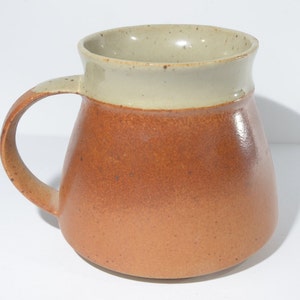 May include: A brown and beige ceramic mug with a wide, rounded base and a handle. The mug has a speckled glaze.