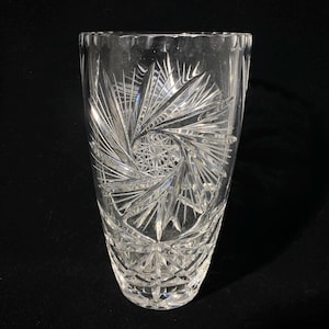 May include: A clear, cut-glass vase with a flared top and intricate starburst and geometric patterns. The vase is cylindrical with a wide opening and a detailed design that refracts light.