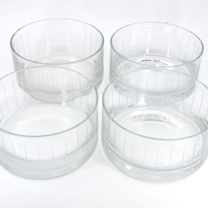 Puede incluir: Cuatro cuencos de vidrio transparente con líneas verticales grabadas. Los cuencos son redondos, de perfil bajo, y están sobre una superficie blanca. El vidrio es transparente, permitiendo ver el fondo blanco.