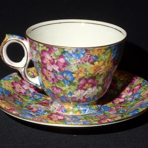 Puede incluir: Un juego de taza y platillo de té floral con un patrón floral rosa, azul y amarillo. La taza tiene un borde dorado y un asa delicada.