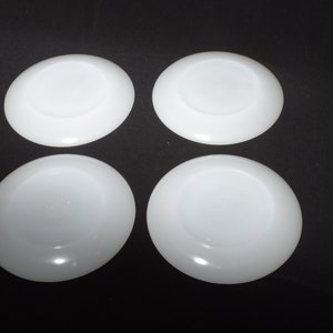 May include: Four white ceramic saucers, each with a smooth, slightly raised center. The saucers are arranged in a square pattern on a black background.