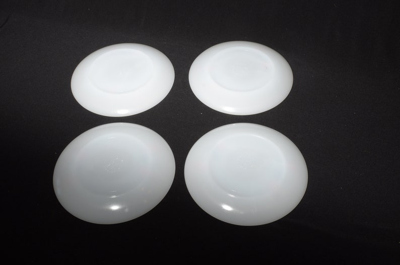 May include: Four white ceramic saucers, each with a smooth, slightly raised center. The saucers are arranged in a square pattern on a black background.