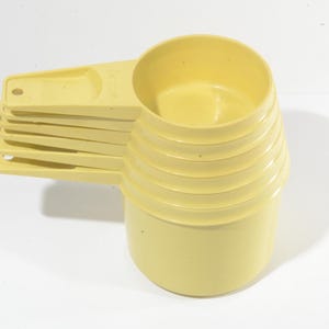 Puede incluir: Un conjunto de seis tazas y cucharas medidoras de color amarillo pálido. Las tazas están apiladas, con los mangos de las cucharas extendidos hacia un lado. El juego es de plástico y está diseñado para uso en la cocina.
