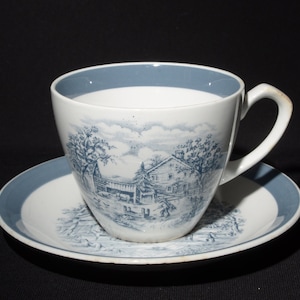 May include: A blue and white teacup and saucer set featuring a country scene design. The cup has a blue rim and the saucer has a blue band around the edge.