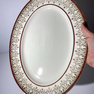 May include: An oval serving platter with a cream-colored center and a decorative border. The border features a maroon band with gold filigree designs. The platter is held by a hand.