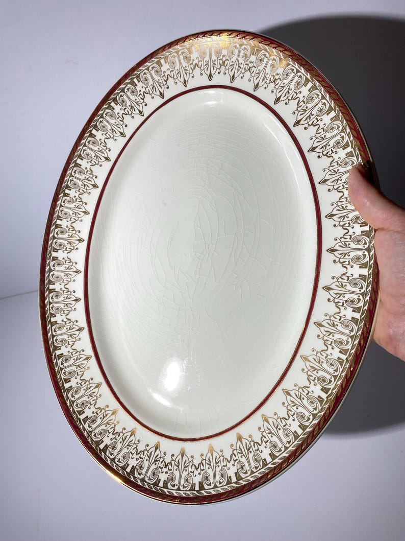 May include: An oval serving platter with a cream-colored center and a decorative border. The border features a maroon band with gold filigree designs. The platter is held by a hand.