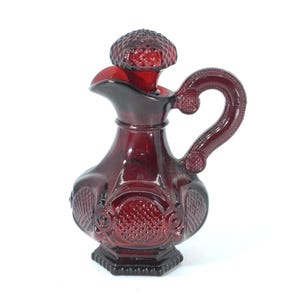 Puede incluir: Una jarra de vidrio rojo rubí con asa y tapón. La jarra tiene un diseño decorativo y un pico vertedor. El tapón tiene forma de hongo. La jarra mide aproximadamente 15 cm de altura.