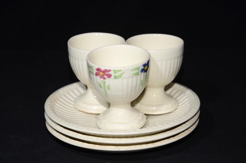 May include: Three white ceramic egg cups with floral designs, sitting on a white ceramic saucer with a fluted edge.