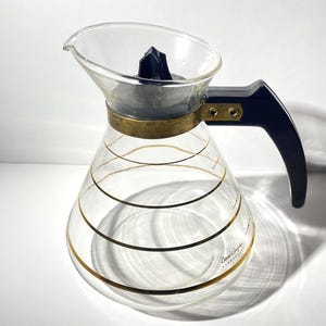 Könnte beinhalten: Eine klare Glas-Kaffeekanne mit einem schwarzen Kunststoffgriff und -deckel. Die Kanne hat goldene Streifen um den Körper und den Text "Bodum Colombia" auf dem Boden.