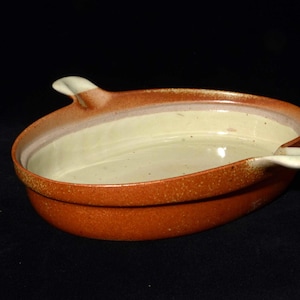 May include: A ceramic oval baking dish with a speckled orange exterior and a cream-colored interior. The dish has two small, white handles and is set against a black background. This stoneware dish is ideal for baking and serving.