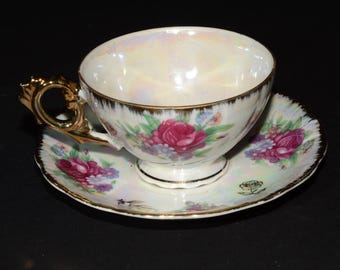 Puede incluir: Un juego de taza y platillo de té blanco con un diseño floral. La taza y el platillo están decorados con flores rosas, rojas y moradas. La taza tiene un borde dorado y un asa dorada. El platillo tiene un borde dorado.