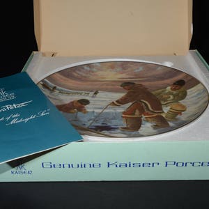 May include: A blue and white porcelain plate with a painting of people in traditional clothing standing on a snowy landscape. The plate is in a box with the words "Genuine Kaiser Porcelain" printed on the front. The text on the plate reads "Nori Peter "People of the Midnight Sun"