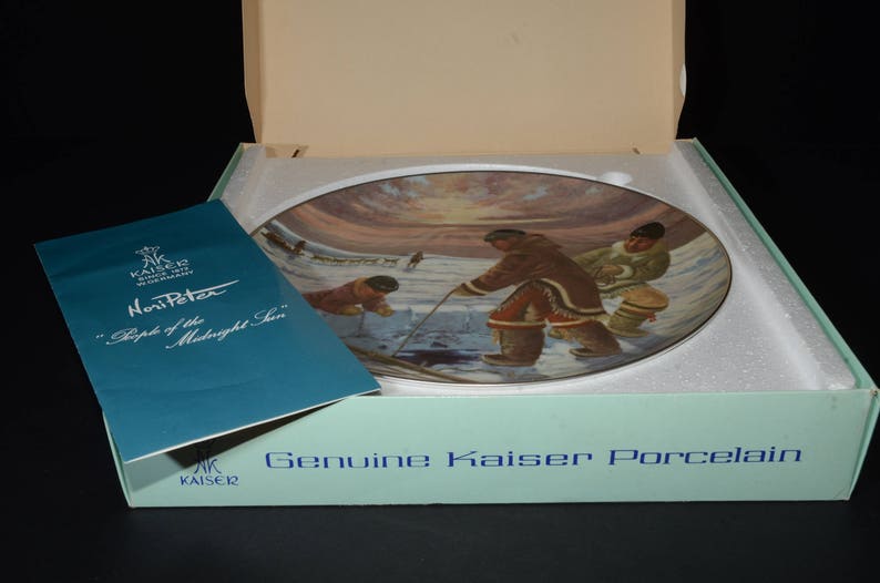 May include: A blue and white porcelain plate with a painting of people in traditional clothing standing on a snowy landscape. The plate is in a box with the words "Genuine Kaiser Porcelain" printed on the front. The text on the plate reads "Nori Peter "People of the Midnight Sun"