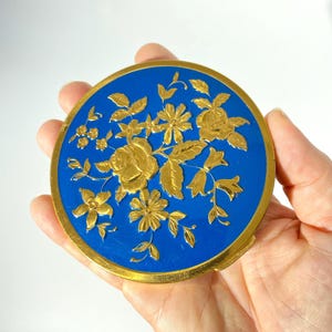 May include: A round compact mirror featuring a vibrant blue enamel surface decorated with an intricate, raised gold-tone floral pattern. The circular case is framed by a polished gold-tone metal edge, creating a distinct contrast between the blue and gold.