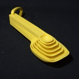 May include: A set of six yellow measuring spoons, nested together with a small loop for hanging. The spoons are made of plastic and vary in size, with a rounded bowl shape. The set is on a black background.