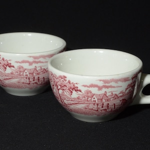May include: Two white ceramic teacups with pink floral designs. The cups have a rounded shape and a handle on the right side. The floral design features a cottage and trees in a landscape setting.