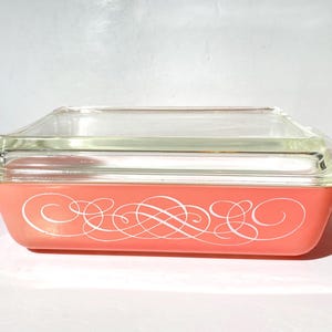 Könnte beinhalten: Rechteckige, korallenfarbene Pyrex-Auflaufform mit einem Deckel aus klarem Glas. Die Form hat ein weißes, verziertes Scroll-Design auf der Vorderseite. Der Deckel liegt bündig auf der Form.