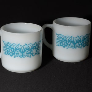 May include: Two white ceramic mugs with a turquoise floral pattern. Each mug has a curved handle and a cylindrical shape. The floral design is a repeating pattern of flowers and leaves around the middle of the mug.