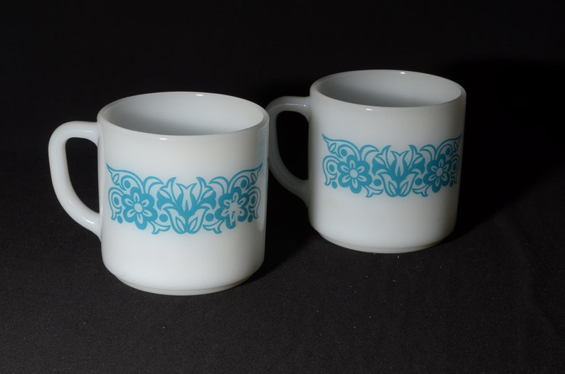 May include: Two white ceramic mugs with a turquoise floral pattern. Each mug has a curved handle and a cylindrical shape. The floral design is a repeating pattern of flowers and leaves around the middle of the mug.