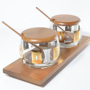 Könnte beinhalten: Ein Set aus zwei Glasbehältern mit goldenen und weißen geometrischen Mustern und Holzdeckeln und -löffeln, auf einem Holztablett präsentiert.