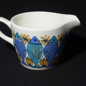 Può includere: Un brocca per la panna in ceramica bianca con un design di pesci blu e gialli. La brocca ha un manico e un beccuccio.