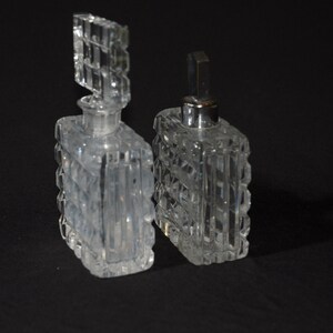May include: Two clear glass perfume bottles with textured surfaces and silver tops. The bottles are square shaped and have a geometric design.