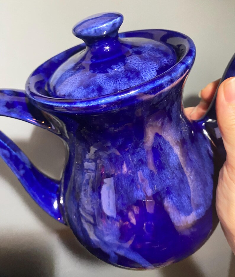 May include: A blue ceramic teapot with a white glaze. The teapot has a round body and a lid with a knob on top. The teapot is decorated with a swirling pattern of white and blue.