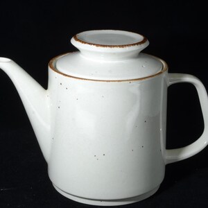 Può includere: Teiera in ceramica bianca con coperchio e manico. La teiera ha un beccuccio e un bordo marrone attorno al coperchio e al bordo. La teiera è su sfondo nero.