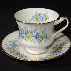 Peut inclure: Une tasse à thé et une soucoupe blanches avec un motif floral de myosotis bleus et de feuilles vertes. La tasse et la soucoupe ont une bordure dorée.
